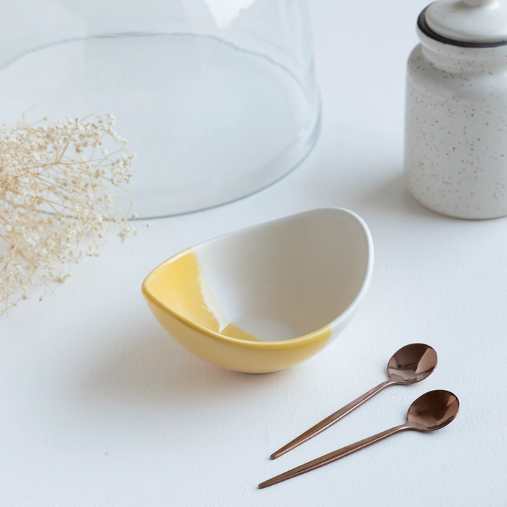 A white and yellow ceramic dessert bowl beside two copper spoons, a ceramic jar on a tabletop.
