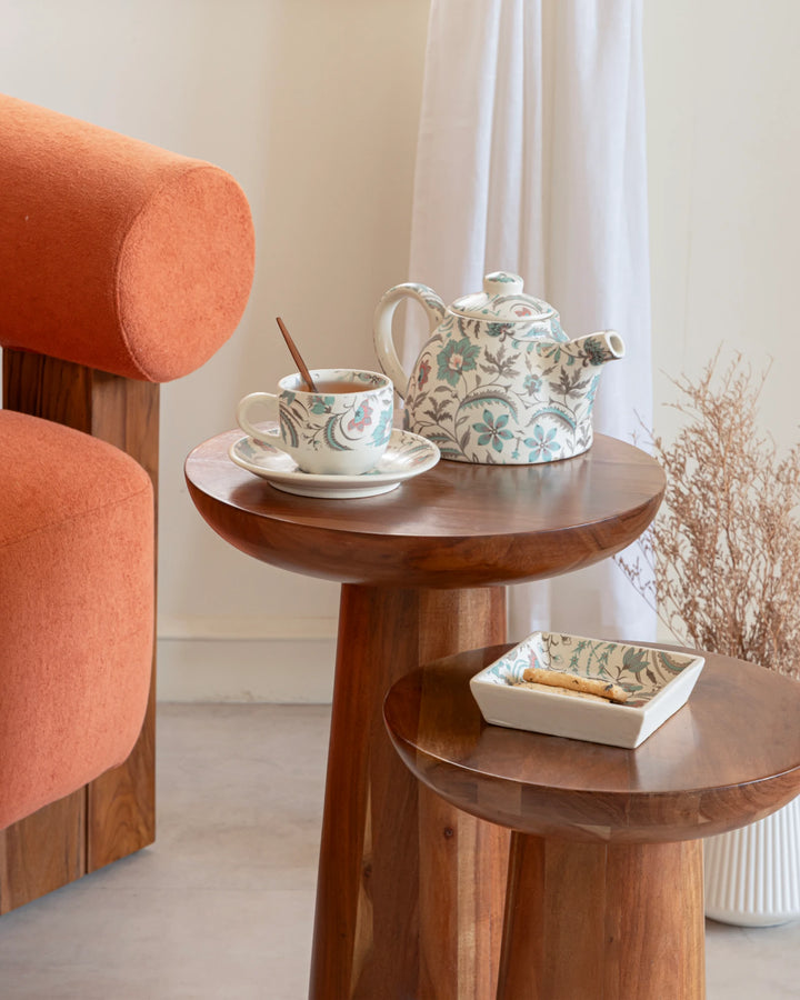 Tea set on a wooden side table with an orange chair and white curtains in the background.