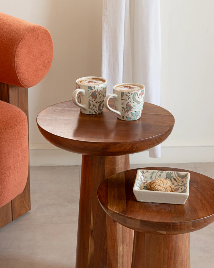Two patterned mugs on a wooden side table with a bowl of cookies, next to an orange armchair.