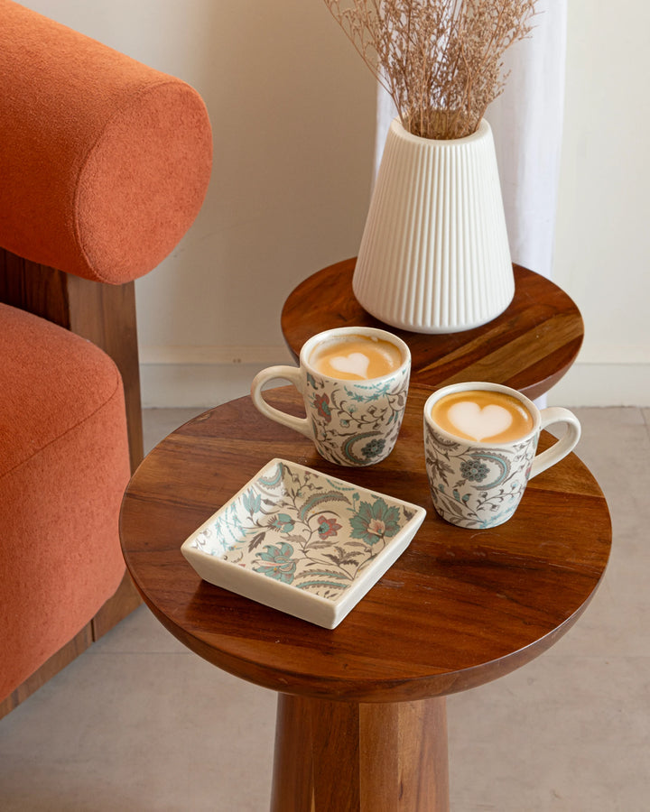 Two mugs with floral patterns on a wooden table next to an orange chair.