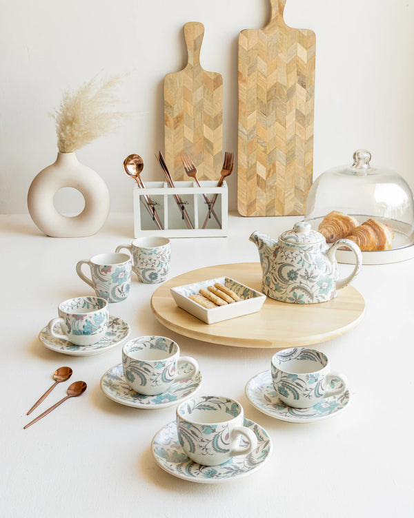 Luxury blue floral tea set with matching teapot and cups, arranged on wooden tray. Elevate your tea experience with this elegant teaware, perfect for gifting or personal use. Comes with spoons set and charming ceramic mugs, complemented by fresh pastries on a glass cake stand and minimalistic wooden wall decor.