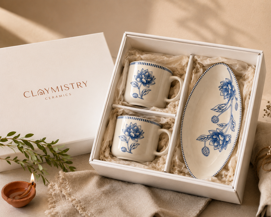 A boxed ceramic gift set from Claymistry Ceramics, featuring two small white mugs with blue floral illustrations and a matching oval plate, displayed in a white gift box with shredded paper. The white branded box lid sits beside the set on a beige fabric surface with soft natural shadows.