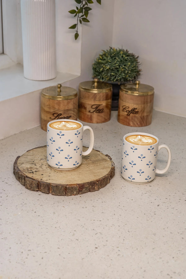 Two mugs with a latte on a wooden coaster, with decorative canisters in the background.