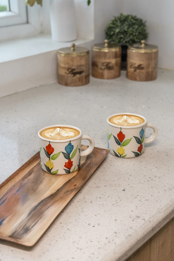 Two ceramic coffee mugs with floral designs on a countertop with decorative jars in the background.