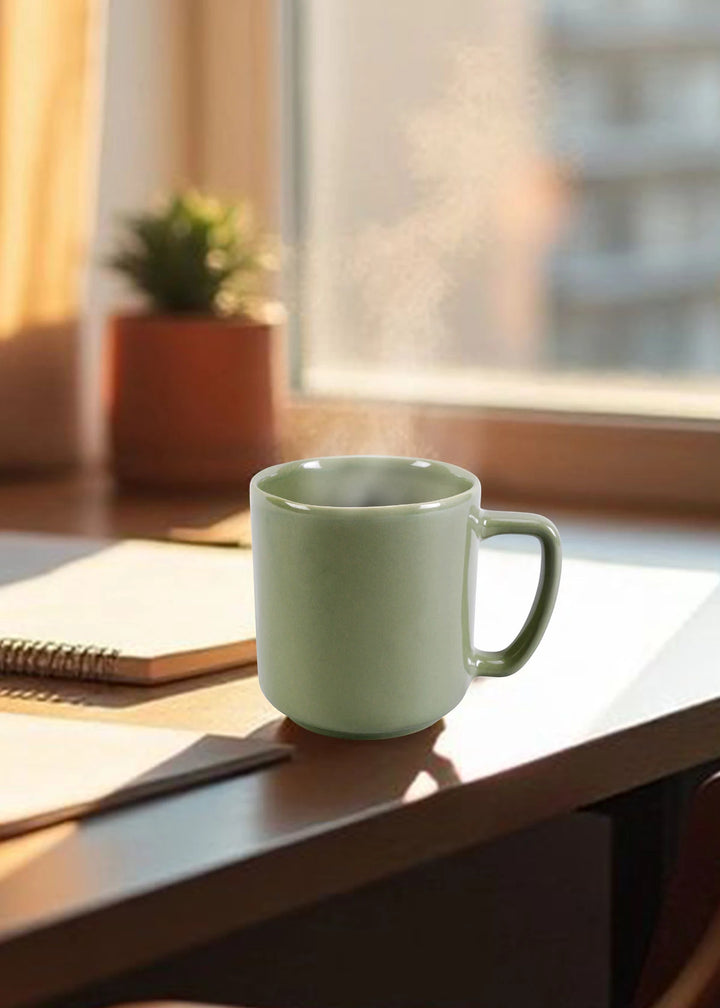 Green mug on a desk with a notebook and plant in the background