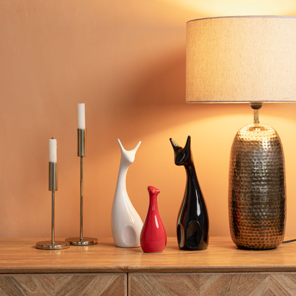 A warm-hued room with candlesticks, 3 ceramic deer-shaped figurines, and a lit lamp on a wooden sideboard