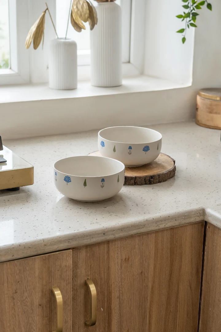 Two The Solitude elegant ceramic dinnerware bowls, adorned with tasteful blue and green tree patterns, displayed on a wooden slice and a speckled kitchen counter, emphasizing their aesthetic appeal as part of artisanal home decor.
