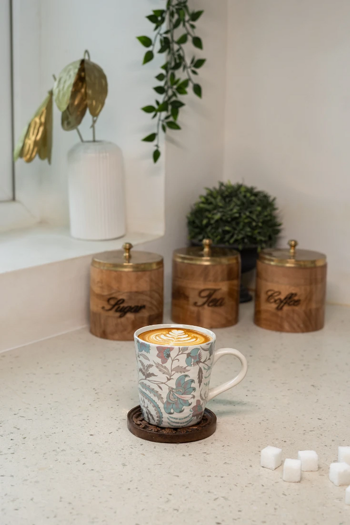Mug with a floral pattern filled with a hot beverage on a countertop with wooden canisters labeled 'Tea', 'Sugar', and 'Coffee'.