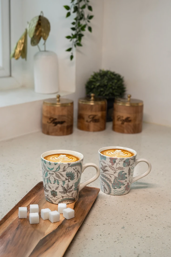 Two floral-patterned mugs with coffee on a wooden tray with sugar cubes, against a neutral background.