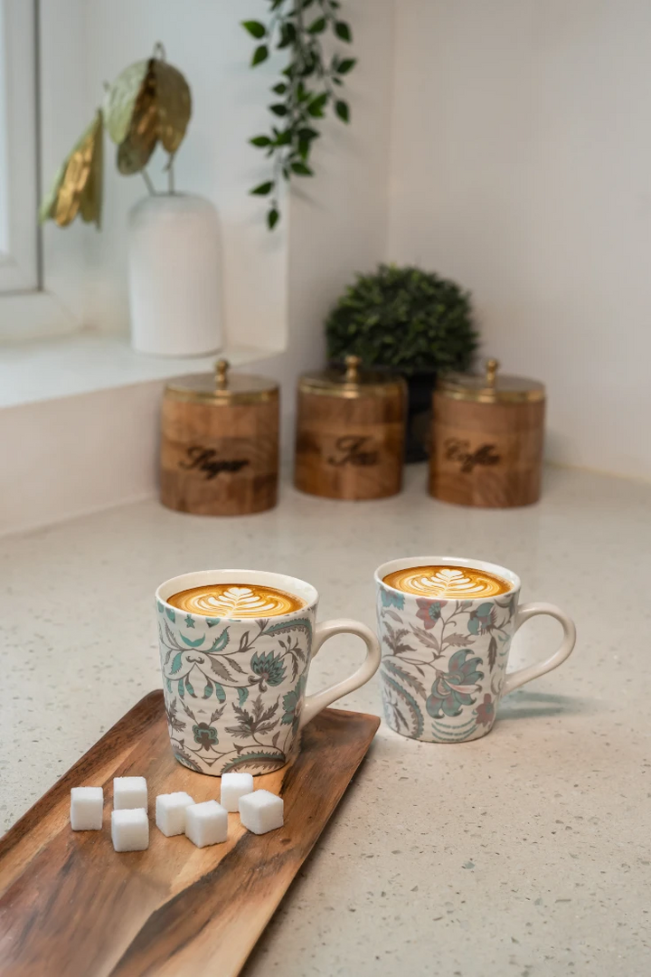 Two floral-patterned mugs with coffee on a wooden tray with sugar cubes, against a neutral background.