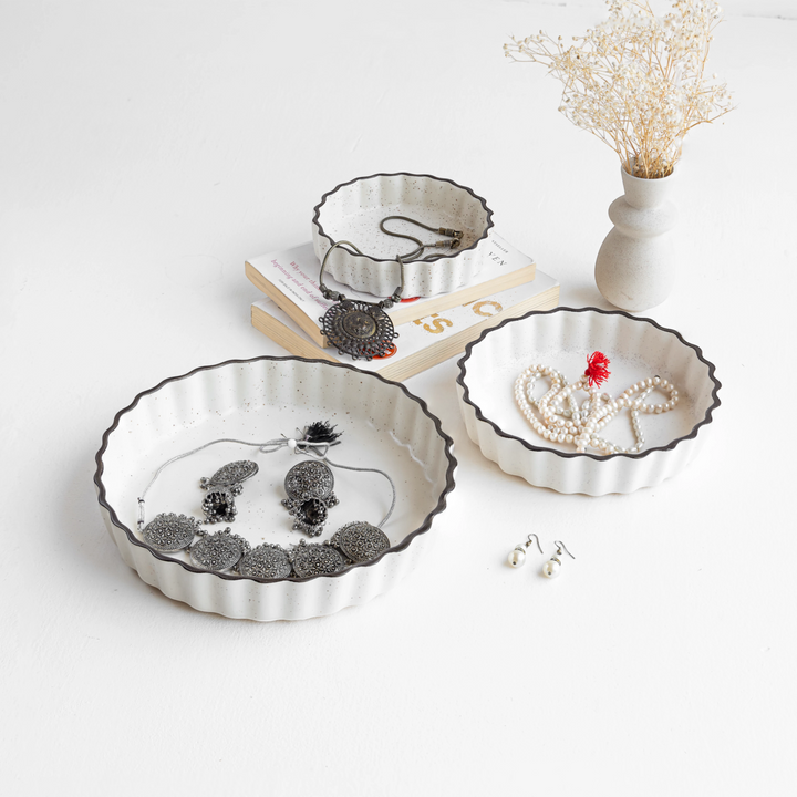 Three scalloped-edge ceramic serving trays holding jewelry on a white surface