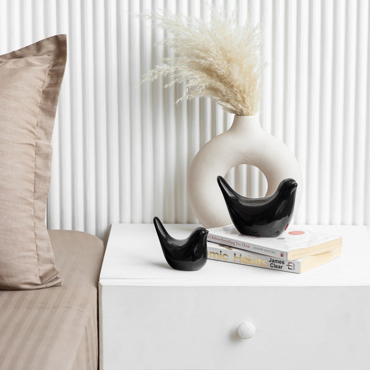 A white ceramic vase placed on books, alongside black bird ceramic figurines on a white cabinet
