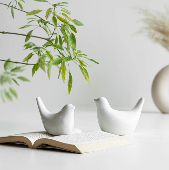 Two white bird ceramic figurines displayed next to an open book on a white surface