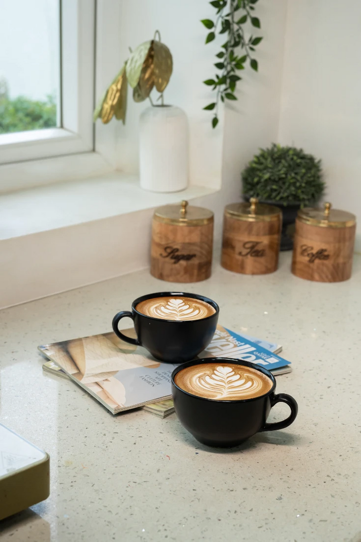 Two black coffee cups with latte art on a light surface, with decorative items in the background.