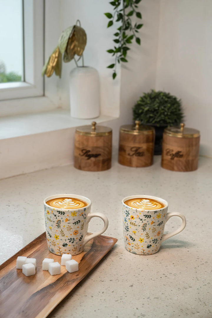 Two floral-patterned mugs with latte art on a light surface, with decorative canisters and a plant in the background.