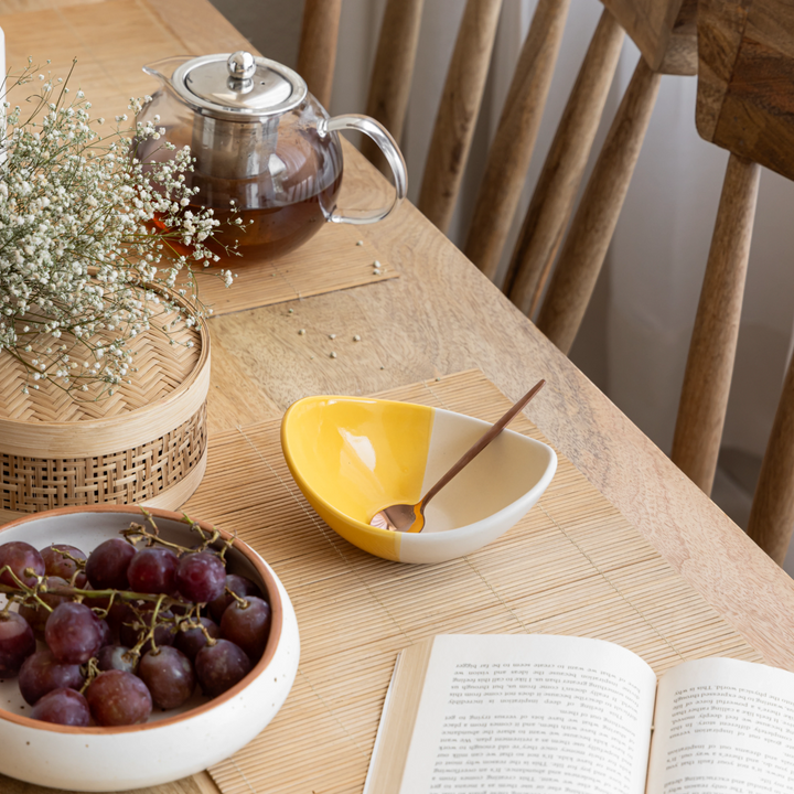 A cozy table scene featuring a teapot, yellow and white ceramic dessert bowl with a spoon, grapes, and a book