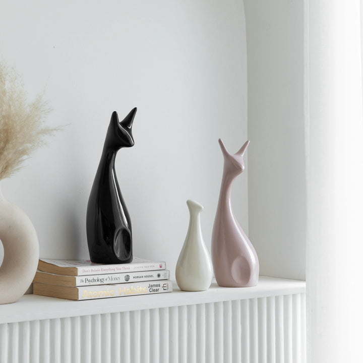 A set of three ceramic figurines named Dashing Deer Figurine, showcasing black, white, and blush pink designs, elegantly styled on a white shelf with books, adding charm to ceramic home decor.