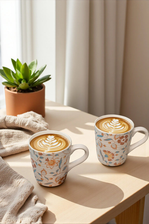 Two floral-patterned coffee mugs with latte art on a wooden surface with a plant and towel in the background.