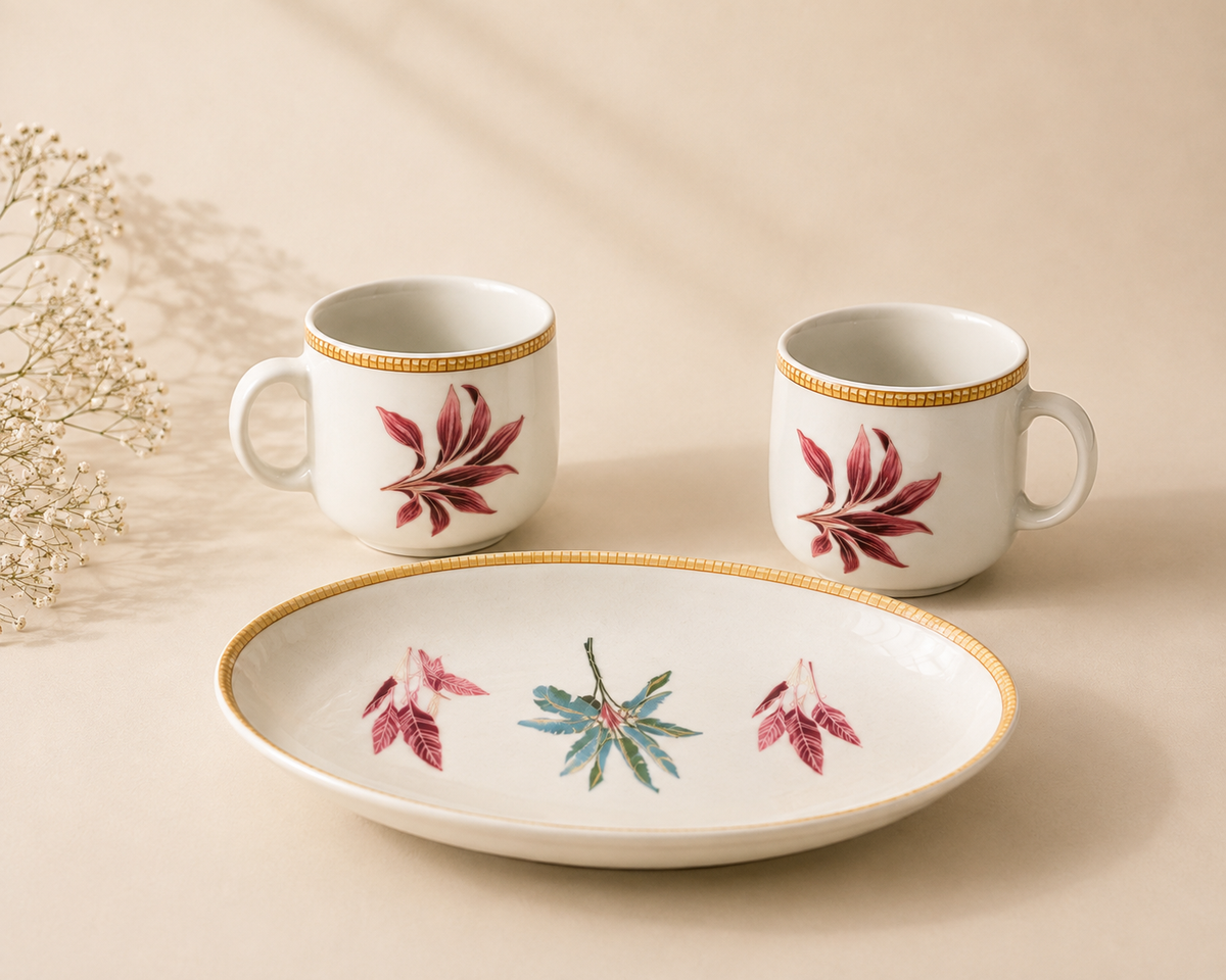 A ceramic tableware set featuring two white handled cups and a matching oval platter, decorated with red and green botanical leaf motifs and gold trim, set against a warm beige background with dried flowers.