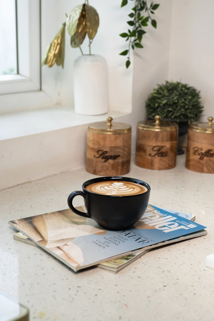 Black mug with cappuccino on a magazine next to wooden containers labeled 'Sugar', 'Tea', and 'Coffee' on a light-colored surface.