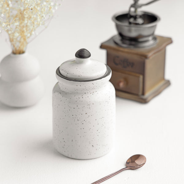 A white ceramic jar with a lid, a spoon, and a coffee grinder placed on a white surface