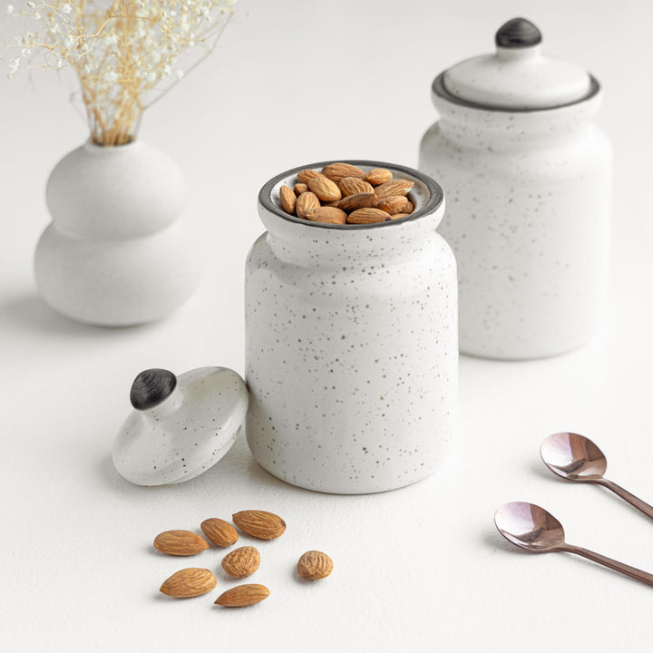 Two white ceramic jars with lids, one filled with almonds, arranged on a white surface