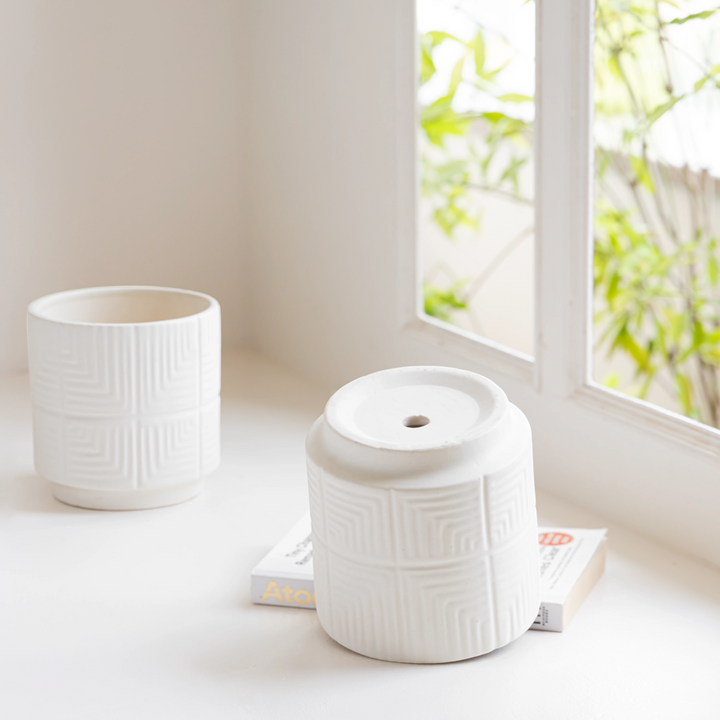 Two textured white ceramic planters sit on a minimalist table near a window